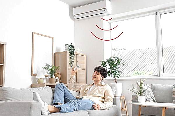 Junger Erwachsener liegt fröhlich auf Sofa und bedient eine Klimaanlage per Fernbedienung